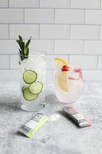 A clean, high-contrast product hero shot featuring two electrolyte drink preparations with fresh fruit garnishes set against a white subway tile background.